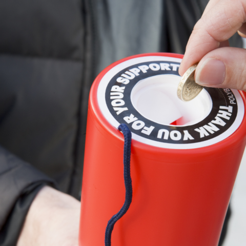 The image shows a close-up of a red charity collection tin being held by a person's hand.