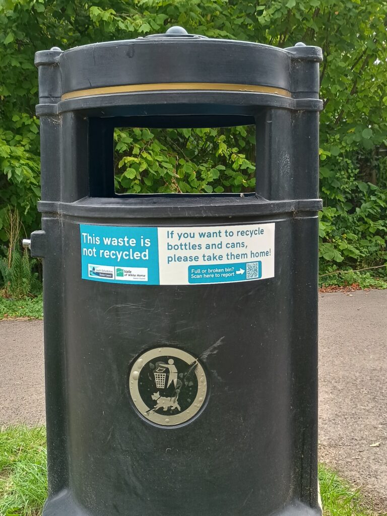 The image shows a black public litter bin with a sign on the front. The sign is split into two sections:

The left section, on a light blue background, says "This waste is not recycled." Below this, there are two logos, one for "Vale of White Horse District Council" and another for "South Oxfordshire District Council."

The right section, on a white background, says "If you want to recycle bottles and cans, please take them home!" Below this is a smaller text that says "Full or overflowing bin? Scan to report" next to a QR code.

The bin itself has a small metal emblem with a graphic of a person putting a piece of trash into a bin.