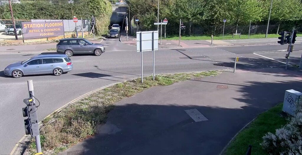 Detailed Description
The view is taken from a slightly elevated perspective, overlooking the junction.

Foreground:

In the immediate lower left, there is a piece of street furniture (possibly a camera, sensor, or utility box) mounted on a pole, with some overgrown weeds and grass around its base.

The foreground also features a mix of pavement/sidewalk and a small, grassy, and slightly neglected divider area with some low-lying plants.

Mid-Ground (Traffic & Junction):

Two cars are visible on the road:

A blue estate/wagon is driving left across the frame.

A dark grey SUV is turning or driving toward the centre of the frame.

The junction itself is controlled by traffic lights, with one set visible on the far right and another small set visible near the tunnel entrance.

A large, blank signpost stands in the middle of a small island separating two lanes.

Various road markings and pedestrian crossing lines are clearly visible on the tarmac.

Background (Tunnel & Advertising):

The dominant feature in the distance is an underpass or tunnel running beneath what looks like a raised embankment, possibly for a railway line. A third, dark car is emerging from this tunnel.

To the left of the tunnel entrance, there is a large, temporary advertising board on an area of land being developed or used for storage. The sign reads:

"STATION FLOORING" in large red letters.

"RETAIL & COMMERCIAL FLOORING" in smaller letters.

The entire area is bordered by fences and dense trees/bushes on the embankment, giving the area a somewhat industrial or infrastructural feel. Street signs are also scattered throughout the background.

The lighting suggests a sunny day with clear shadows. The overall impression is of a functional, busy urban traffic route near a station or major crossing point.