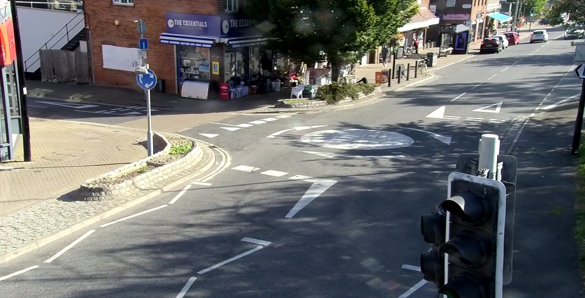 The image is taken from a slightly elevated position, from a cctv camera across the road. Foreground: A set of traffic lights is prominently visible on the right side of the frame. The lights are black and appear to be showing a red signal in this view (though the lamps themselves are dark). The road surface immediately in the foreground has white road markings, including a solid white stop line and dashed lane markings. Mid-Ground: The centre of the image is dominated by a mini-roundabout, indicated by a painted circle with white arrows pointing clockwise around it. Numerous other white dashed lines indicate lanes and pedestrian crossings leading into and out of the roundabout. To the left of the roundabout, there is a raised kerb with a small, tiered planter area. A blue circular sign with white arrows (indicating a mandatory direction of travel) is mounted on a pole near the kerb. Background (Left/Centre): A single-story or ground-floor shop is visible behind the roundabout and raised kerb. The shop has an awning and its signage says 