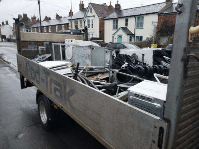 The main subject of the photograph is a trailer filled with scrap metal and large waste items, parked on a wet, paved street. The trailer has high, light grey or silver metal sides and is heavily loaded with junk, including what appears to be part of a washing machine, various metal pipes, black engine parts, and a small white appliance (perhaps a microwave or mini-oven) visible near the foreground. In the background, there is a typical street scene with two-story brick and painted houses, parked cars, and traffic signs.