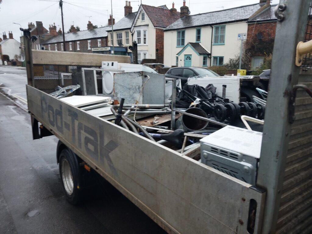 The main subject of the photograph is a trailer filled with scrap metal and large waste items, parked on a wet, paved street. The trailer has high, light grey or silver metal sides and is heavily loaded with junk, including what appears to be part of a washing machine, various metal pipes, black engine parts, and a small white appliance (perhaps a microwave or mini-oven) visible near the foreground. In the background, there is a typical street scene with two-story brick and painted houses, parked cars, and traffic signs.
