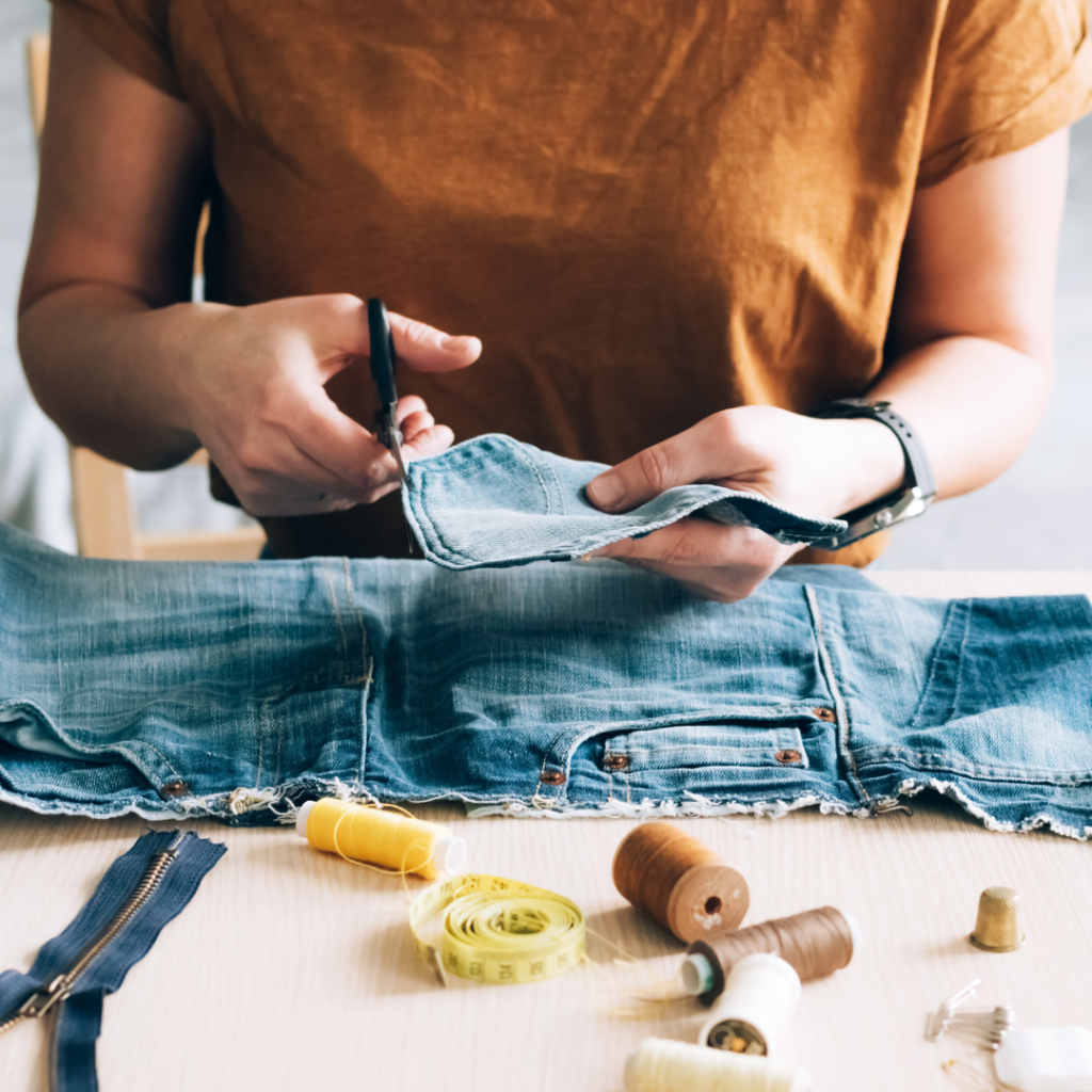 A close-up view of a person using scissors to cut fabric from a pair of denim jeans on a wooden table, surrounded by sewing tools such as spools of thread, a tape measure, a thimble, and a zipper.