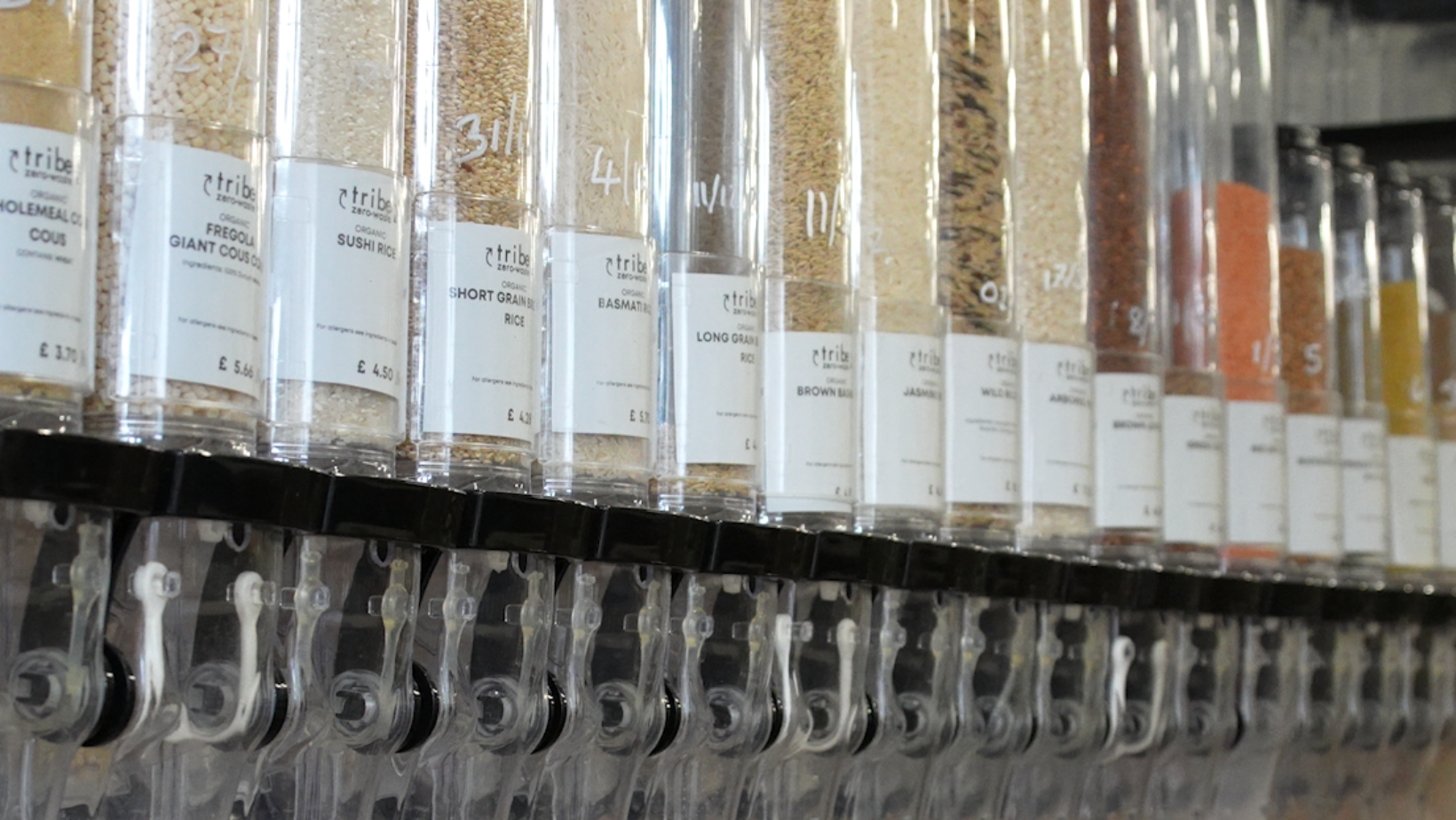 A close-up view of a zero-waste refills station inside a store. Transparent gravity dispenser bins line a wooden shelving unit, filled with various dry grains, pasta, rice, and seeds labeled with product names and prices. Below the main wall dispensers sit glass jars containing seeds and spices, along with a small black sign reminding customers to weigh their empty containers before filling. Additional rows of bulk bins fade into the background on the right.