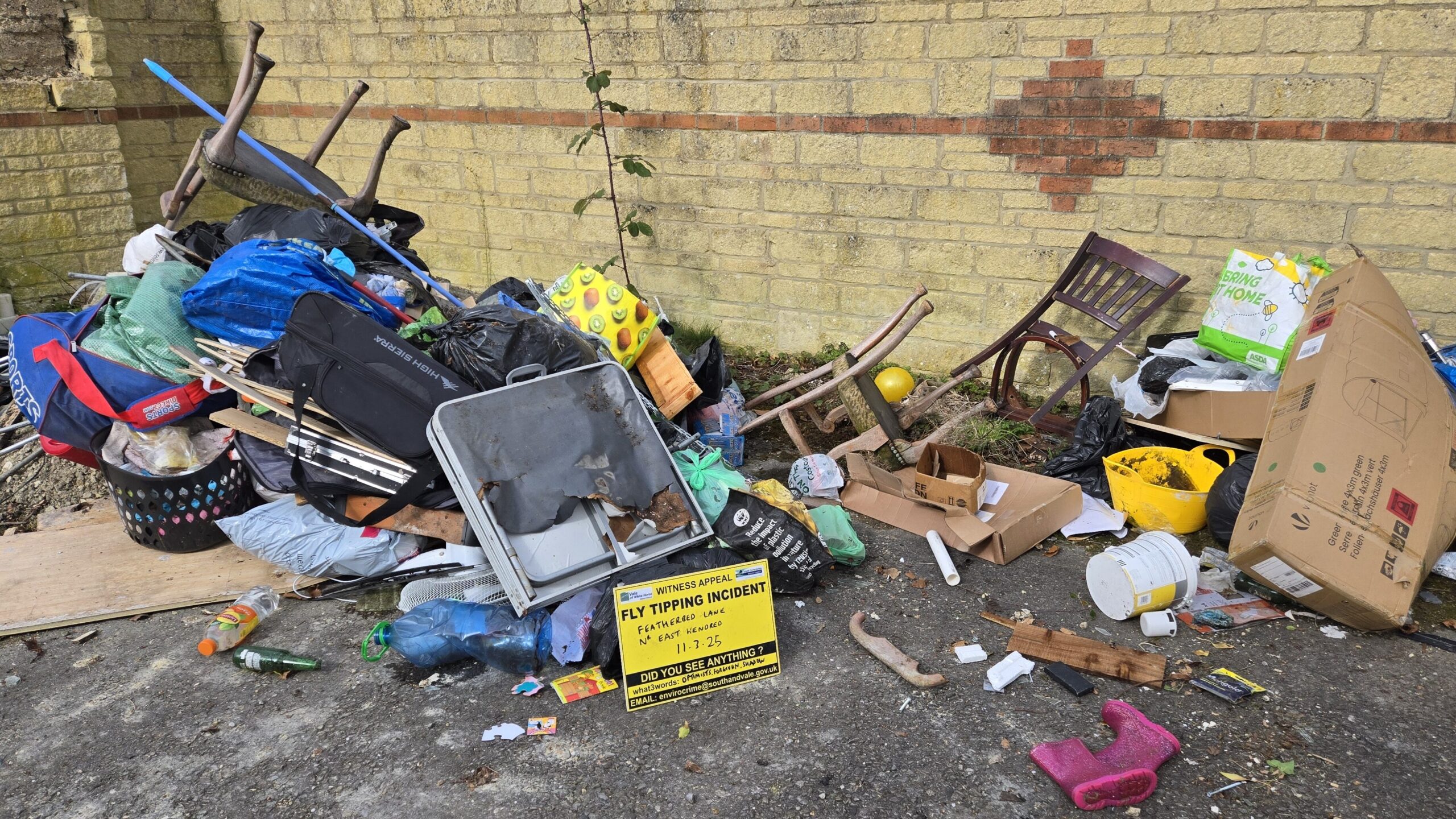 A large pile of various household rubbish and debris, including furniture, plastics, and cardboard boxes, illegally dumped against a pale yellow brick wall. A yellow sign with black text reading "WITNESS APPEAL FLY TIPPING INCIDENT" is partially visible in the foreground.
