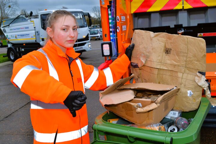 The image is a medium-close shot of a young woman wearing a bright orange, reflective work jacket and black gloves. She is standing outdoors next to a green recycling bin that is overflowing with cardboard boxes and some other plastic and metal waste. The woman is looking directly at the camera with a neutral to slightly serious expression and giving a thumbs-down gesture with her right hand. Her left hand is resting near the large, brown cardboard boxes that are precariously stacked on the bin. She has her blonde hair pulled back. In the background, on a paved area that appears to be a works yard or street, there are several large refuse collection trucks, one of which is white and partially visible on the left, and another with bright orange and yellow reflective hazard stripes on its back is visible on the right. The day is overcast.