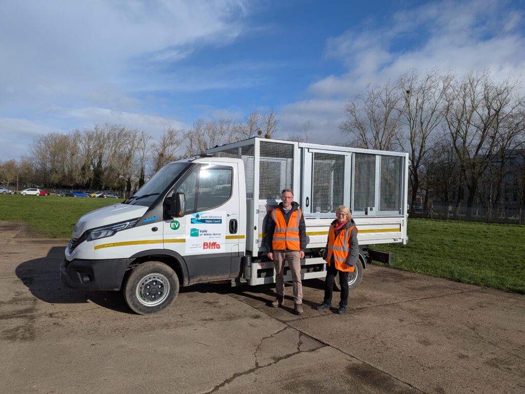 White EV tipper truck on car park with trees and grass in background. In the foreground Cllrs Mark Coleman and Helen Pighills