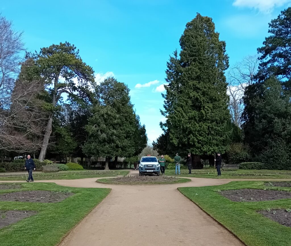 An ornamental gardens fringed with very tall evergreen trees in the background, a van and gardeners working in the foreground