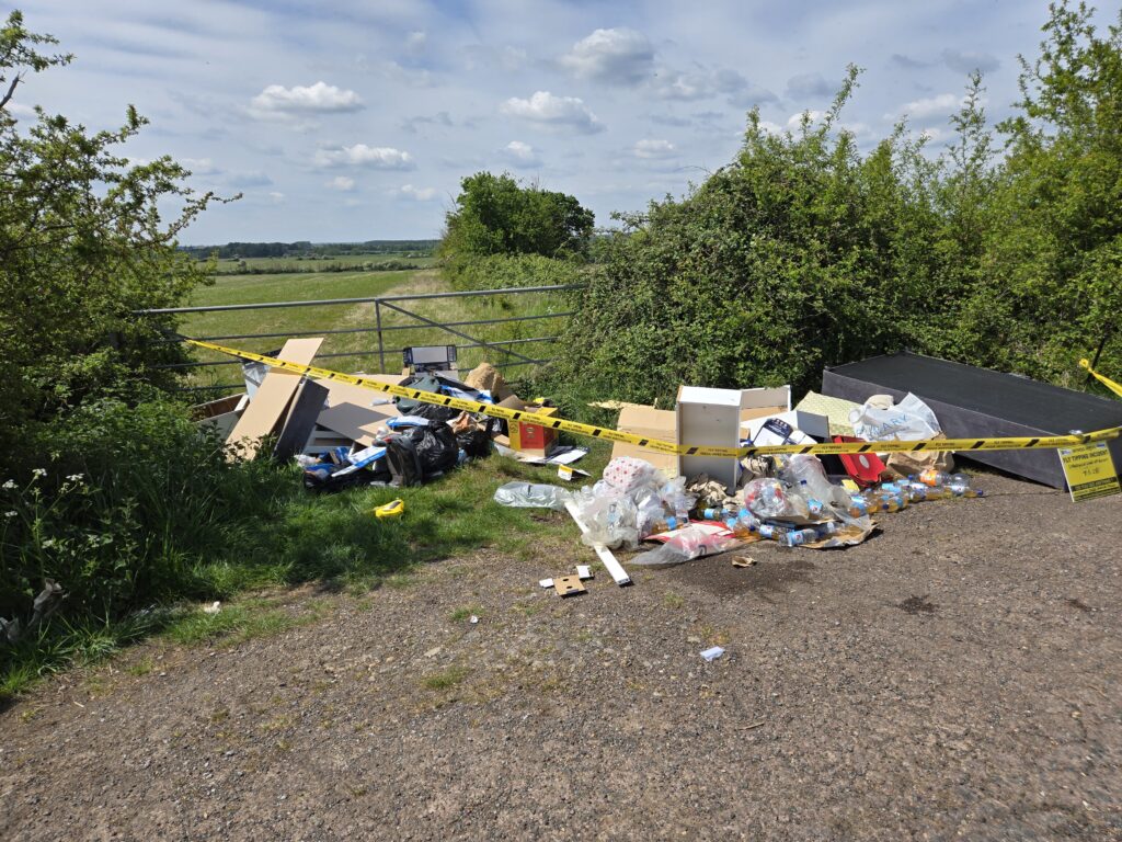 A wide shot of a fly-tipping incident on a rural gravel turnout next to a metal gate and green fields. A large pile of rubbish, including broken white furniture, cardboard boxes, plastic bags, and dozens of plastic bottles containing amber liquid, is cordoned off with yellow "FLY TIPPING UNDER INVESTIGATION" tape. A yellow witness appeal sign is prominently displayed on the right.