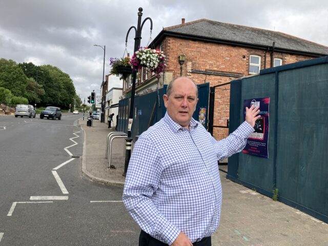 Man stands in front of a development next to a road with traffic