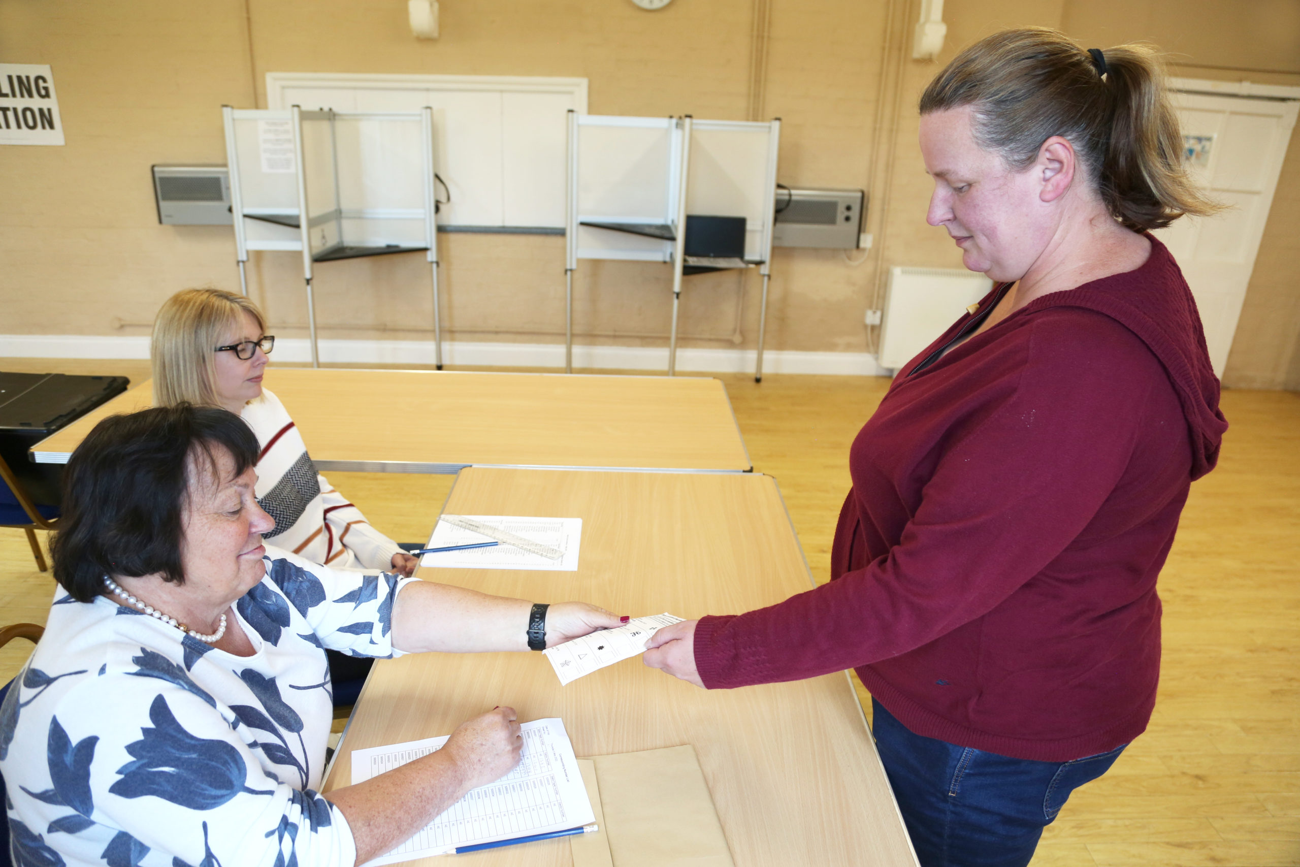 a photo of a lady in a polling station being handed a ballot paper by staff who are sat down at table